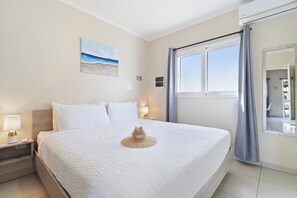 A hotel room with a large bed, a nightstand with a lamp, a painting of a beach, and a window with curtains. - Chic 2BR Apartment w Free Parking near Palm Beach (Noord)