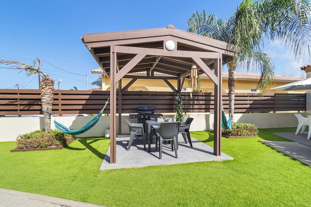 A covered outdoor seating area with a grill, surrounded by artificial grass and palm trees.