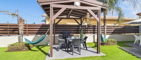 A covered outdoor seating area with a grill, surrounded by artificial grass and palm trees.