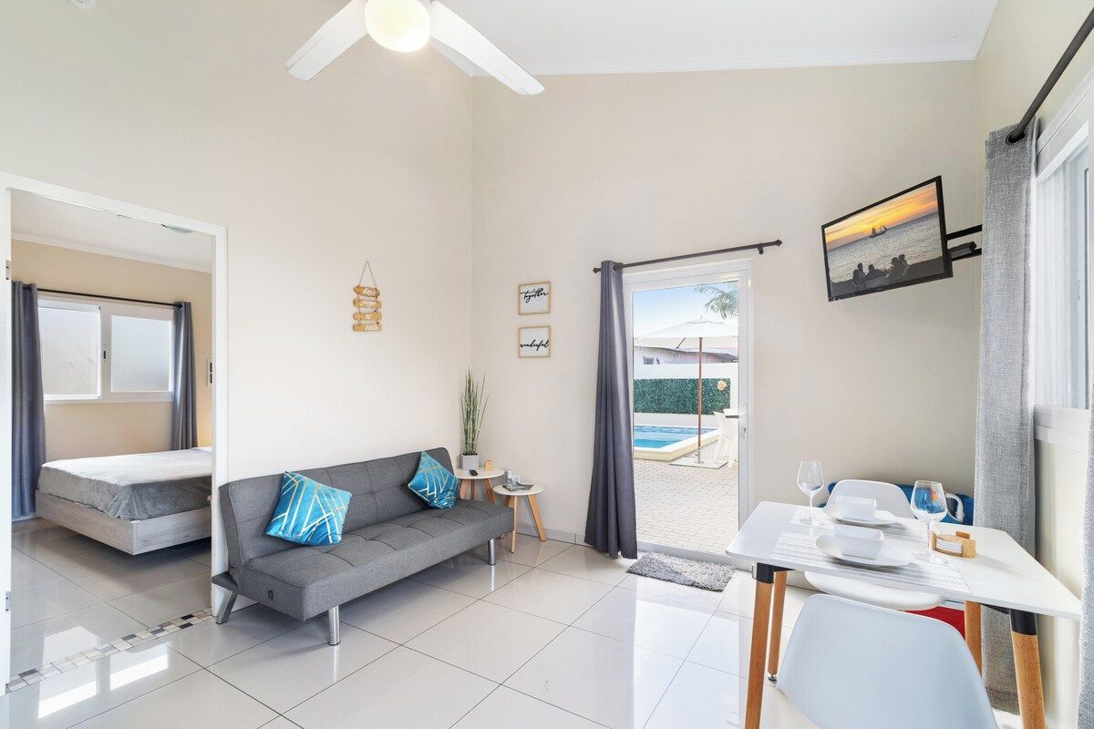 A compact living space with a sofa, a dining table, a small side table, and a view of an outdoor swimming pool.