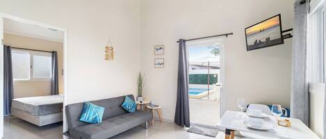 A compact living space with a sofa, a dining table, a small side table, and a view of an outdoor swimming pool.