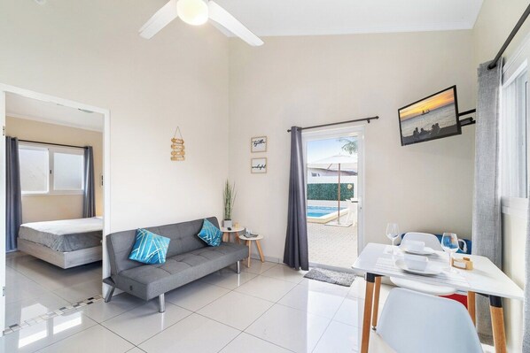 A compact living space with a sofa, a dining table, a small side table, and a view of an outdoor swimming pool.