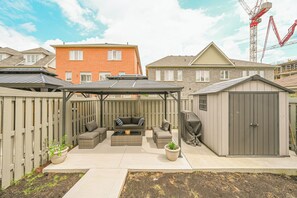 Ein Garten mit einer überdachten Sitzgelegenheit, einem Grill und einem Geräteschuppen.