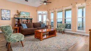 A living room with a brown sofa, a wooden coffee table, and patterned chairs. There is a bookshelf and a painting on the wall. - Bayfront, amazing views, waterside decking, close to everything! – Sunscape (Dauphin Island)