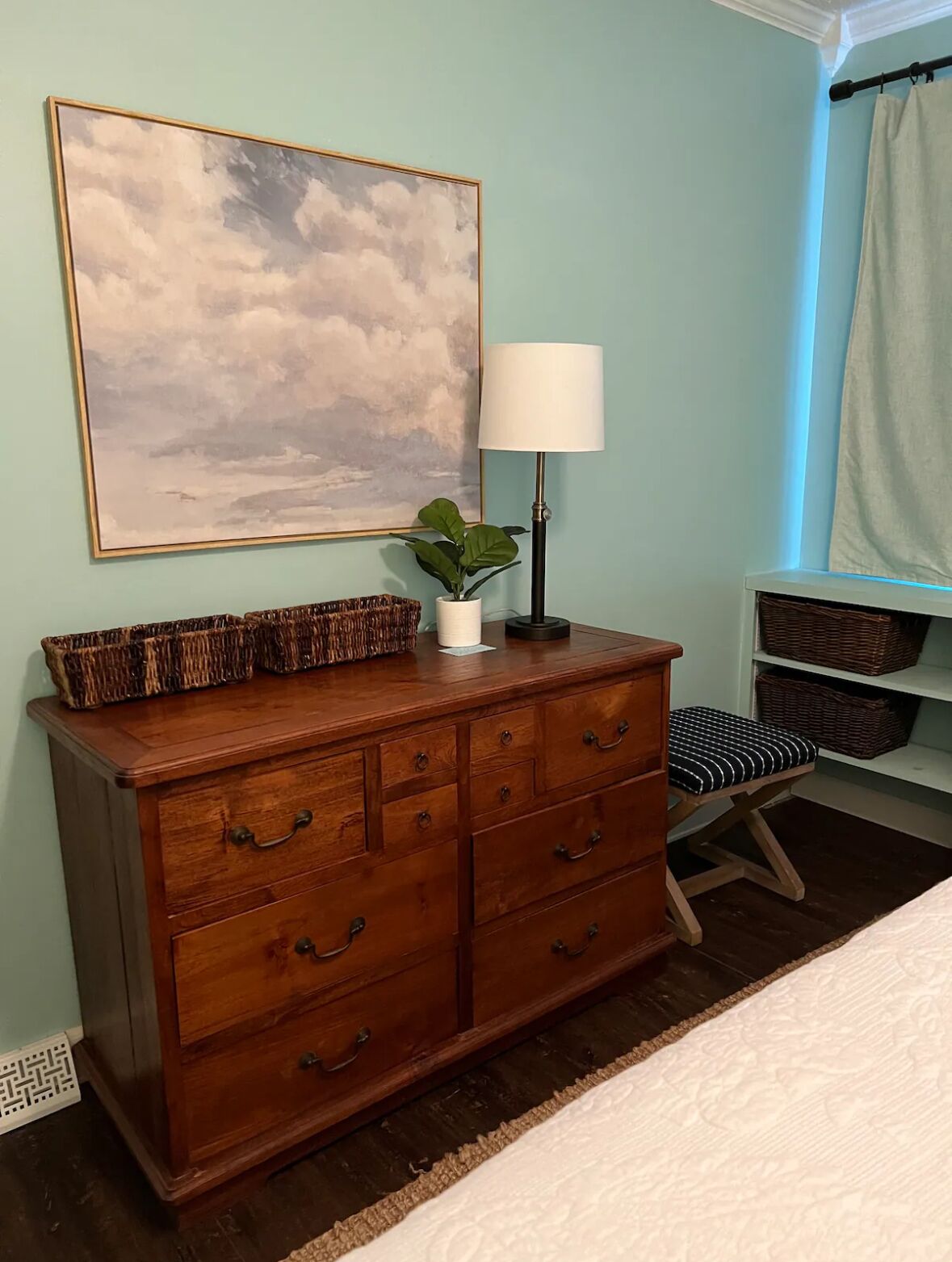 Un dormitorio con un aparador de madera, una mesita de noche, una lámpara, una planta y un cuadro grande en la pared.