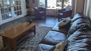 Un salon avec un canapé en cuir, une table basse en bois et un ventilateur de plafond. Une grande fenêtre offre une vue sur l’extérieur.