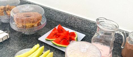 Una mesa de desayuno con diferentes alimentos y bebidas, incluyendo sandía en rodajas, plátanos y una jarra de bebida rosa.