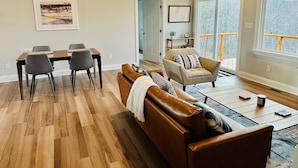 A spacious living room with a wooden floor, a brown leather sofa, a dining table with chairs, and large windows offering a view of trees. - Nature getaway; beautiful Mountain Views, close to rivers, breweries & wineries. (Amherst)