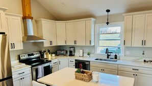 A modern kitchen with white cabinets, stainless steel appliances, a central island with chairs, and a window providing natural light. - Nature getaway; beautiful Mountain Views, close to rivers, breweries & wineries. (Amherst)
