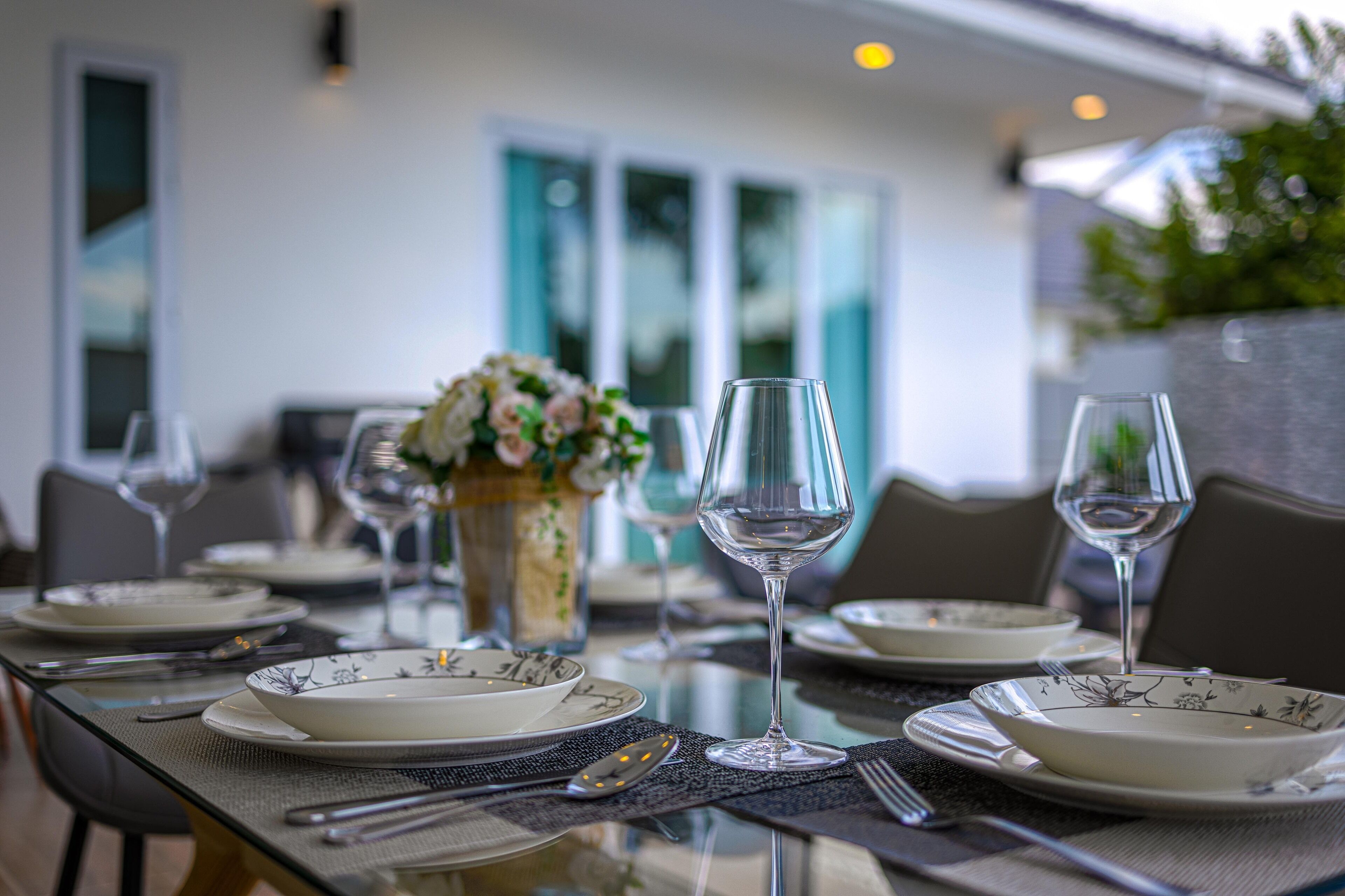 A dining table set with plates, glasses, and cutlery, with a vase of flowers in the background.