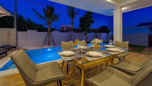 Un espace extérieur aménagé pour les repas, avec une piscine, entouré de palmiers et bordé d’un mur blanc.