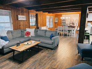 Uma sala de estar acolhedora com um sofá seccional cinza, uma mesa de centro de madeira e uma área de jantar com mesa e cadeiras brancas.
