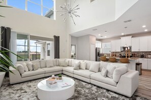 A modern living room with a large L-shaped sectional sofa, a round coffee table, and a kitchen in the background. - Last Minute Disney! Private Pool, Game Room & Themed Bedrooms. Book Now! #56087 (Orlando)