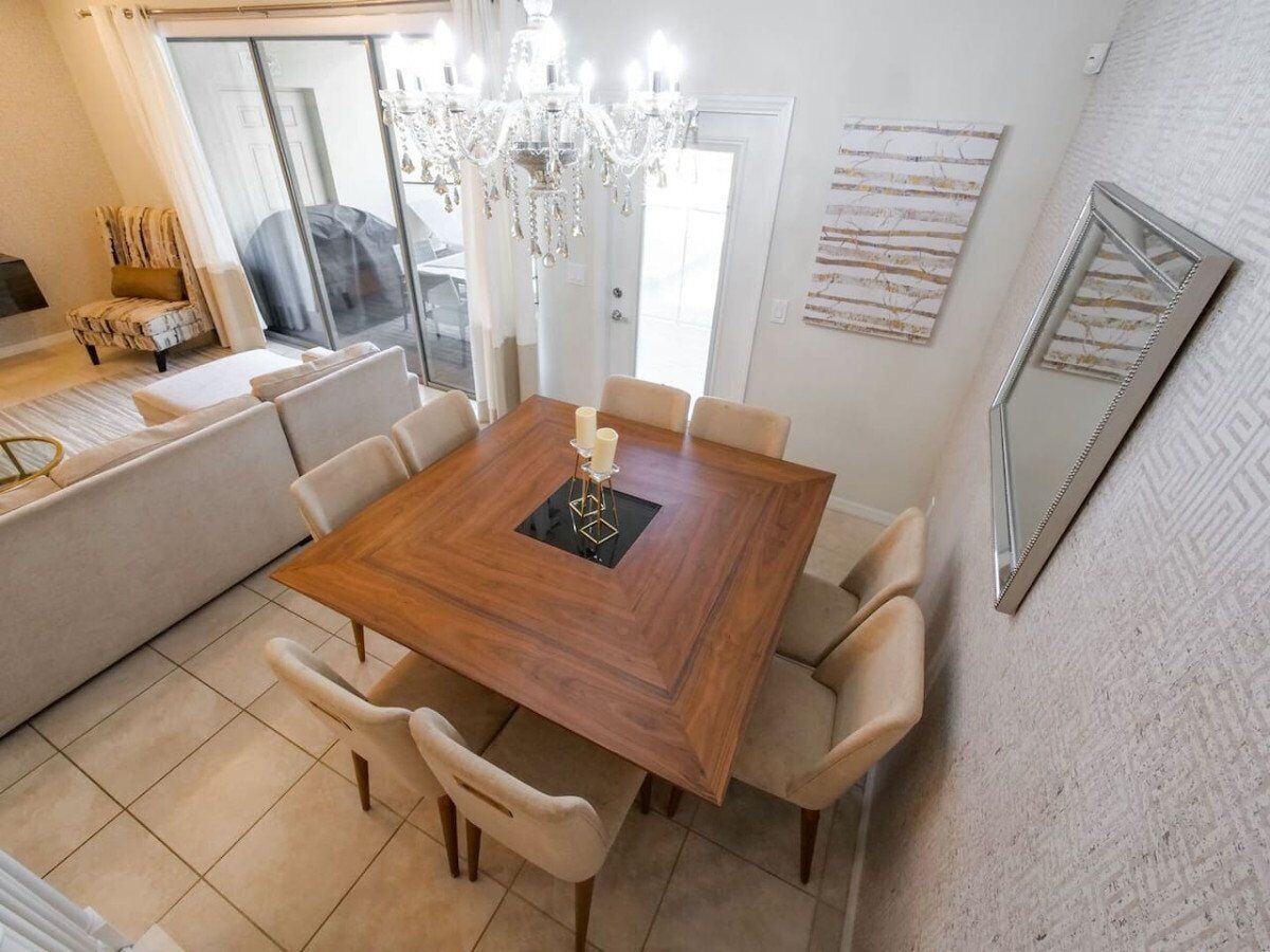A dining area with a wooden table and six chairs, a chandelier, and a large window with curtains.