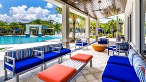 Ein überdachter Außenbereich mit Sitzgelegenheiten, blau-orangenen Kissen, einem Pool und im Hintergrund Palmen.