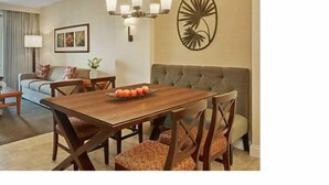 A dining area with a wooden table, four chairs, and a fruit bowl. There is a sofa and a lamp in the background.