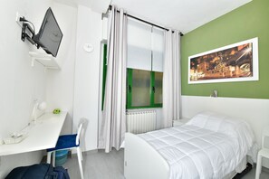 Ein Schlafzimmer mit Bett, Schreibtisch mit Stuhl, Fenster mit Vorhängen, Heizkörper und einer Wanduhr.
