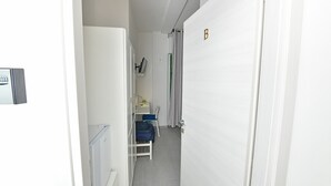 Un couloir étroit avec une porte blanche portant l’inscription « B », un petit coin salon avec une chaise et une table, et un radiateur sur le côté gauche.