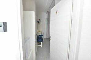 Ein schmaler Flur mit einer weißen Tür, auf der 'B' steht, ein kleiner Sitzbereich mit einem Stuhl und einem Tisch sowie ein Heizkörper links.