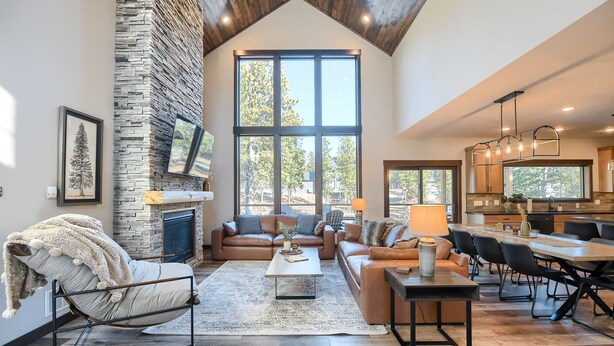 A spacious living room with a stone fireplace, a large window offering a view of trees, a cozy seating area with a sofa and chairs, and a dining area with a table and chairs. - Incredible New 3200+ Sq. Ft. Lodge w/ Arcades, Shuffleboard, Hot Tub, Paved! (Lead)