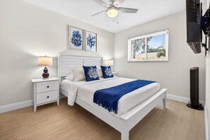 Un dormitorio con una cama blanca, dos mesitas de noche, un ventilador de techo y una ventana con persianas.