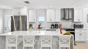 Una cucina moderna con mobili bianchi, un frigorifero in acciaio inossidabile, un'isola centrale con sgabelli da bar e un'ampia finestra che permette l'illuminazione naturale.