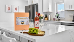 Una cucina moderna con mobili bianchi, un frigorifero in acciaio inossidabile e un piano di lavoro in marmo. Sul piano di lavoro, una bottiglia di Barefoot Pink Moscato, una ciotola di uva e un vassoio di cracker.