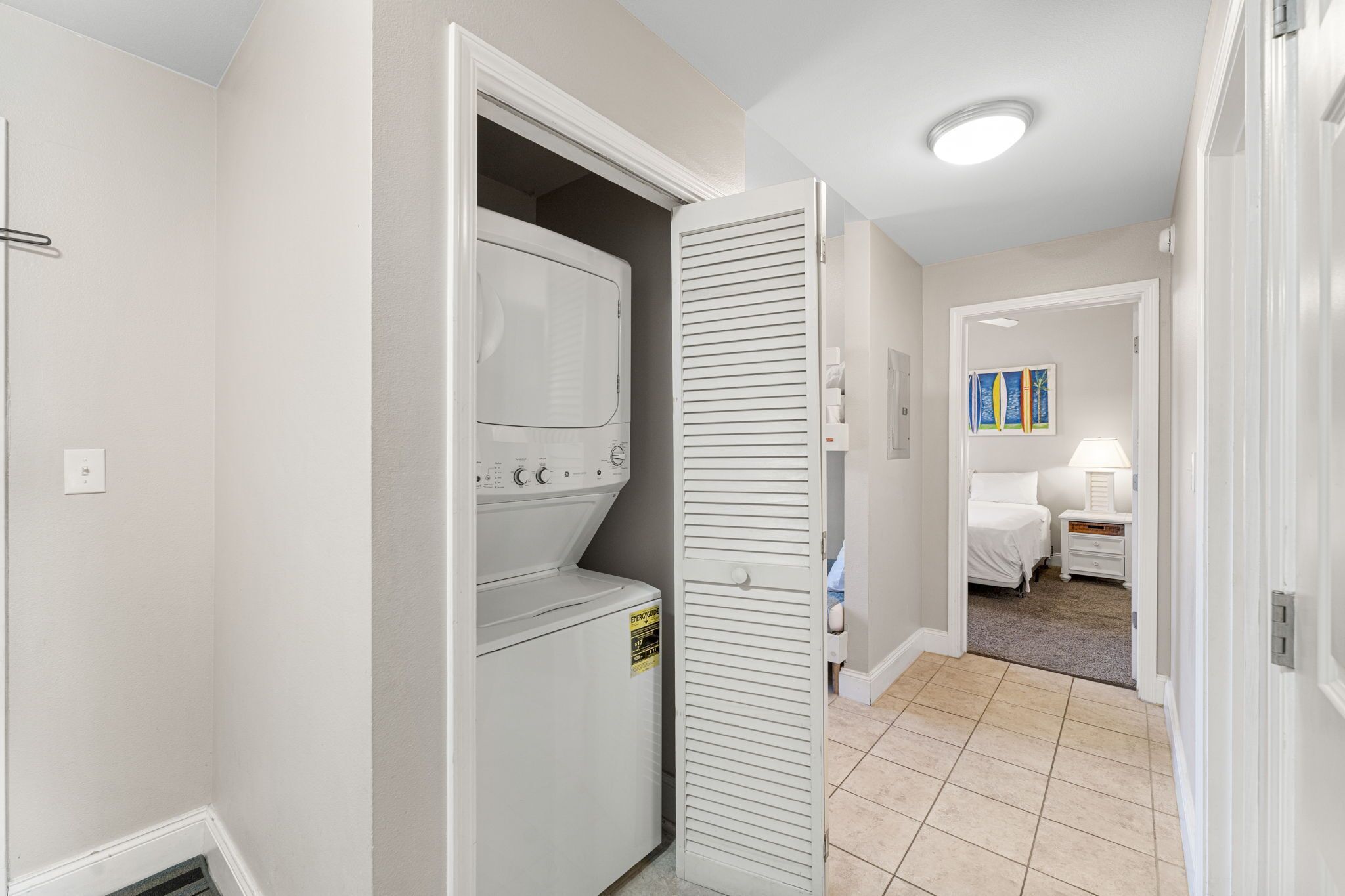 A laundry room with a washer and dryer, a closed closet, and a bedroom with a bed and nightstand.