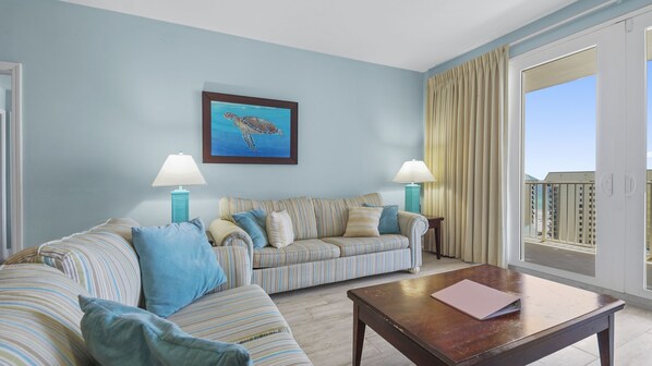 A living room with a striped sofa, two side tables with lamps, a framed painting of a turtle, and a view through a large window. - Waterfront condo w/ daily attraction tickets (Panama City Beach)