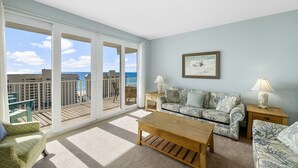 A spacious living room with a large sliding glass door leading to a balcony with a view of the ocean. There is a sofa, a coffee table, and two side tables with lamps.
