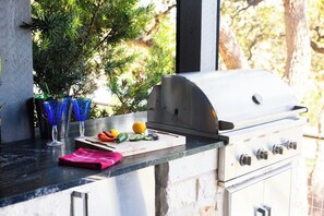 An outdoor kitchen with a grill, a cutting board with vegetables, and a window with a view of trees. - Lola's Lakefront: 3-Bed Treehouse with Porch, Fire Pit & Year-Round Fun (Spicewood)