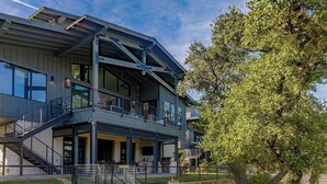 A modern two-story house with a balcony, a stone retaining wall, and a large tree. - Lola's Lakefront: 3-Bed Treehouse with Porch, Fire Pit & Year-Round Fun (Spicewood)