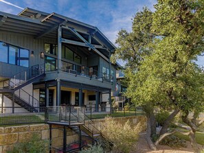 A modern two-story house with a balcony, a stone retaining wall, and a large tree. - Lola's Lakefront: 3-Bed Treehouse with Porch, Fire Pit & Year-Round Fun (Spicewood)
