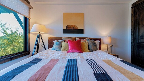 A bedroom with a large bed, a headboard, a window, and a painting on the wall. - Lola's Lakefront: 3-Bed Treehouse with Porch, Fire Pit & Year-Round Fun (Spicewood)