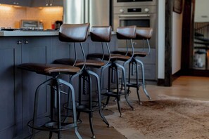 A row of bar stools with metal frames and leather seats, positioned in a kitchen area with cabinets and appliances. - Lola's Lakefront: 3-Bed Treehouse with Porch, Fire Pit & Year-Round Fun (Spicewood)
