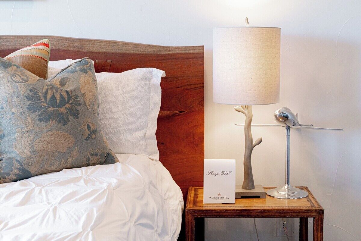 Una cama bien hecha con edredón blanco y almohadones decorativos, una mesita de noche con lámpara y cartel, y un cabecero de madera.