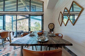 A dining area with a wooden table, chairs, and a view through large windows. - Lola's Lakefront: 3-Bed Treehouse with Porch, Fire Pit & Year-Round Fun (Spicewood)