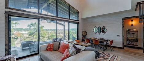 A modern living room with a large window, a sofa, a dining area, and a kitchen visible through an open door.