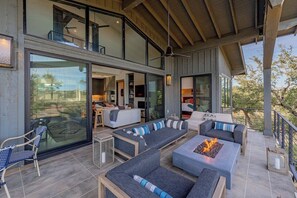 A spacious outdoor living area with modern furniture, a fire pit, and a view of the interior. - Lola's Lakefront: 3-Bed Treehouse with Porch, Fire Pit & Year-Round Fun (Spicewood)
