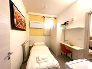 Een kleine, compacte kamer met een enkel bed, een bureau, een stoel, een radiator en een wandairconditioner.