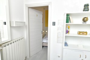Un couloir avec un radiateur blanc, une porte donnant sur une chambre, et une étagère blanche contenant des livres et un globe terrestre.