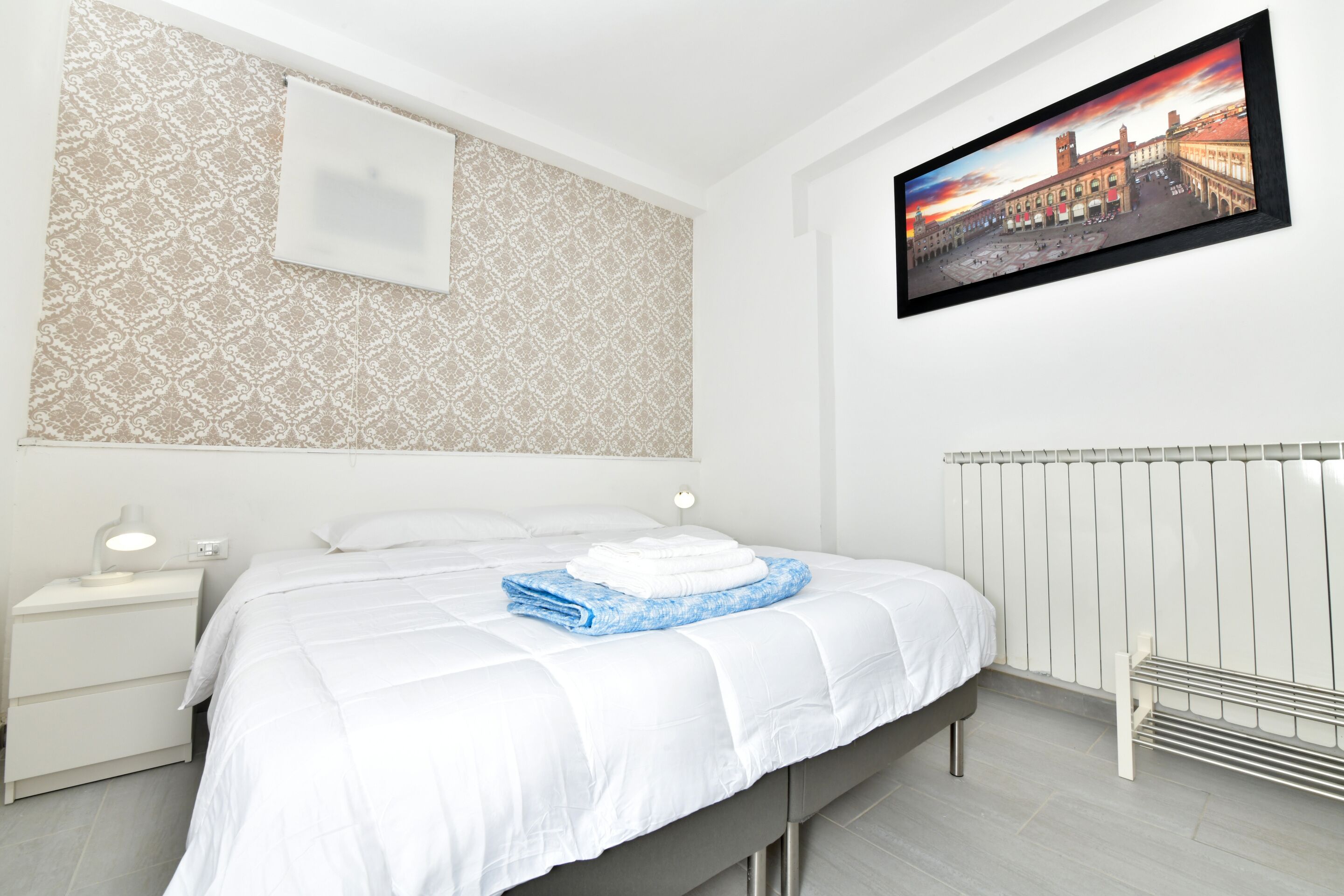 Une chambre d’hôtel avec un lit, une table de chevet, un tableau accroché au mur et un radiateur.
