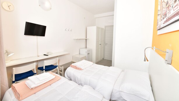 Une chambre d’hôtel avec deux lits, un bureau avec un ordinateur, une télévision et une salle de bain avec un mur orange.
