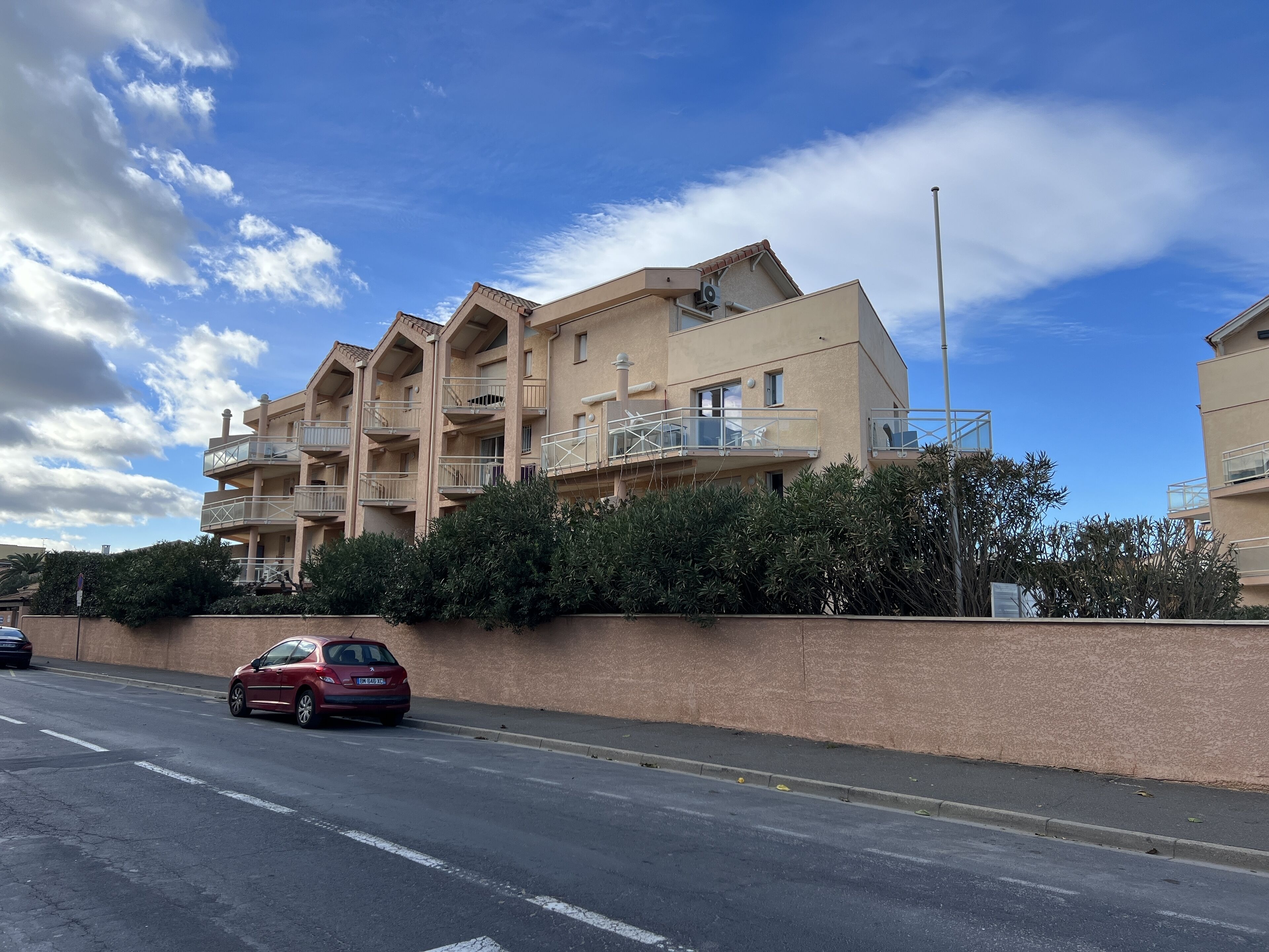 A multi-story residential building with balconies, a parking area, and a clear sky.