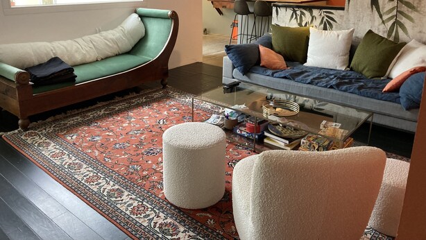 Una sala de estar con un sofá verde, un sofá azul y una mesa de centro de vidrio. Hay una alfombra estampada en el suelo.