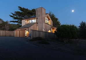 A modern wooden house with a prominent central chimney, large windows, and a well-lit exterior at dusk. - Hawk Haven | Ocean Views, Hot Tub & Cozy Fireplace (Sea Ranch)