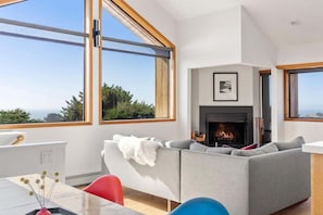 A modern living room with a fireplace, large windows, and a view of the outdoors. - Hawk Haven | Ocean Views, Hot Tub & Cozy Fireplace (Sea Ranch)