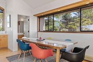 A dining area with a long table, four chairs, and a window with a view of trees. - Hawk Haven | Ocean Views, Hot Tub & Cozy Fireplace (Sea Ranch)