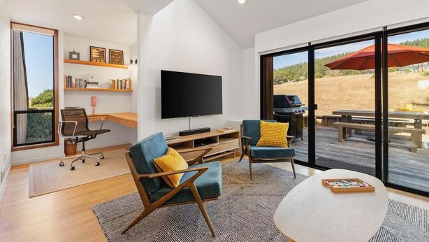 A modern living room with a flat-screen TV, comfortable seating, a wooden coffee table, and a large sliding glass door leading to an outdoor deck.
