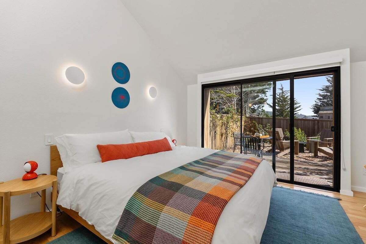 Un dormitorio con cama, mesita de noche, manta colorida y puerta corrediza de vidrio que da a un espacio exterior.